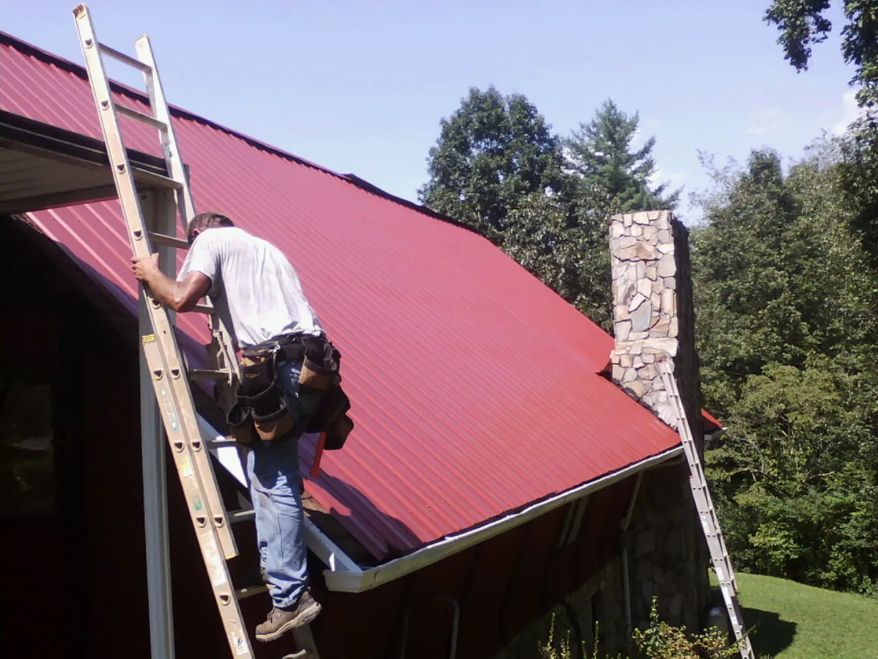 Roofer on a ladder beside a red metal roof and stone chimney — Soffit & Fascia Repair by Roofing Brothers in Glenwood